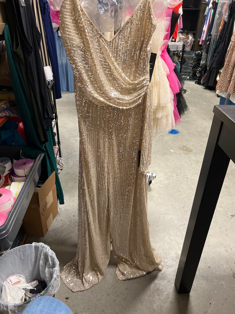 A floor-length champagne gold sequined gown displayed on a dress form in what appears to be a retail or storage room with clothing racks and shelves visible in the background.