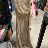 A floor-length champagne gold sequined gown displayed on a dress form in what appears to be a retail or storage room with clothing racks and shelves visible in the background.