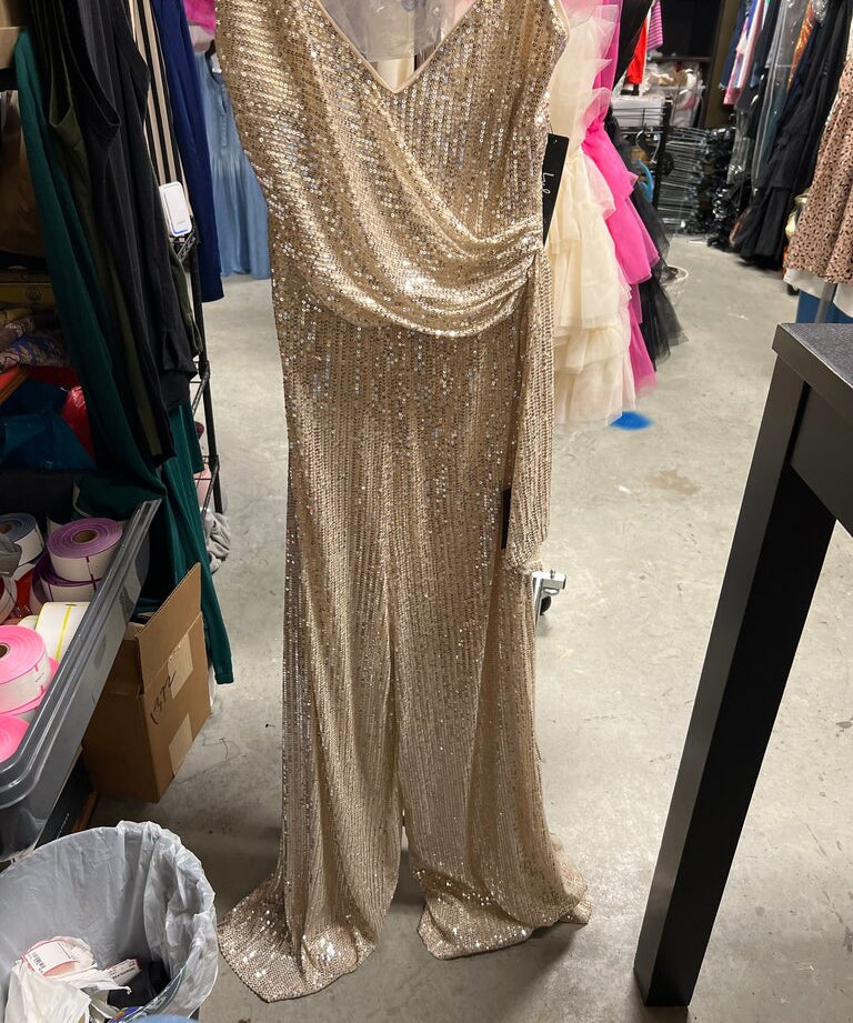 A floor-length champagne gold sequined gown displayed on a dress form in what appears to be a retail or storage room with clothing racks and shelves visible in the background.